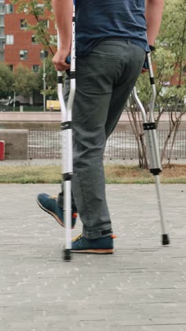 A man barely hobbles on one leg with crutches. Adult male walks slowly down the street, leaning on two crutches. Adult disabled person walking in the park during the day. Close-up shot