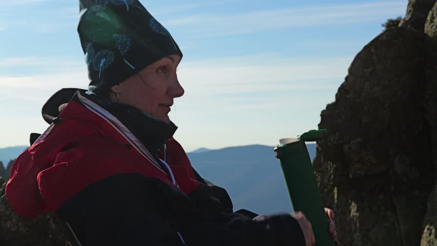 Close-up of a girl in a red-and-black windbreaker drinking green thermos tea, sitting on a rocky ledge against a background of mountain landscape and blue sky. Side view