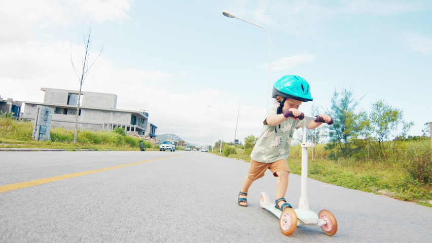 Toddler boy learns to ride a scooter and falls. His mother comes and calms him down. Boy falls from a scooter during learning