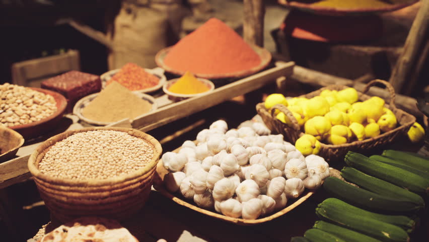 Stalls filled with colorful spices, fresh garlic, lemons, and vibrant vegetables create a lively atmosphere in a traditional African market bustling with activity.