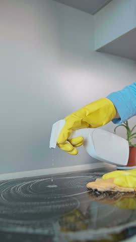 A woman in an apron cleaning the kitchen stove with cleaning products. Wet cleaning with disinfectants kills bacteria left after cooking.
