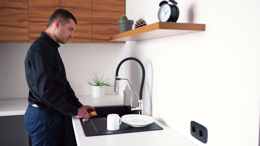 Young man stand in fron of sink and opens fauset for washing dishes. Take yellow sponge and start cleaning white plates. Washing dishes after lunch or taking meal.