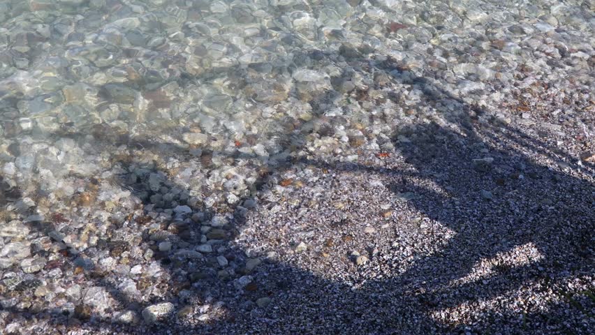 Shadows dance over the crystal-clear waters of Lake Garda in Sirmione, reflecting sunlight on smooth pebbles in Italy