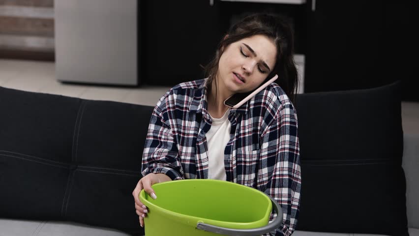 Young woman in kitchen during quarantine. Sit on sofa and talking on phone with plumber. Water liking fron ceiling. Girl need help with fixing pipe.