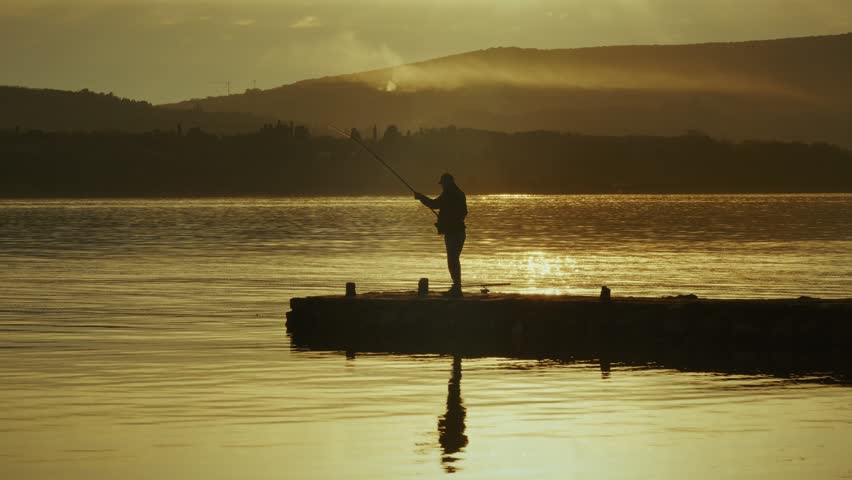 A person enjoys fishing from a dock while the sun sets over serene waters and distant hills.