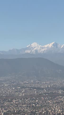 View of Kathmandu’s unplanned urban sprawl encroaching on lower hills, pushing against green spaces. The city’s dense expansion contrasts with distant melting snowcapped mountains, a stark sign of cli