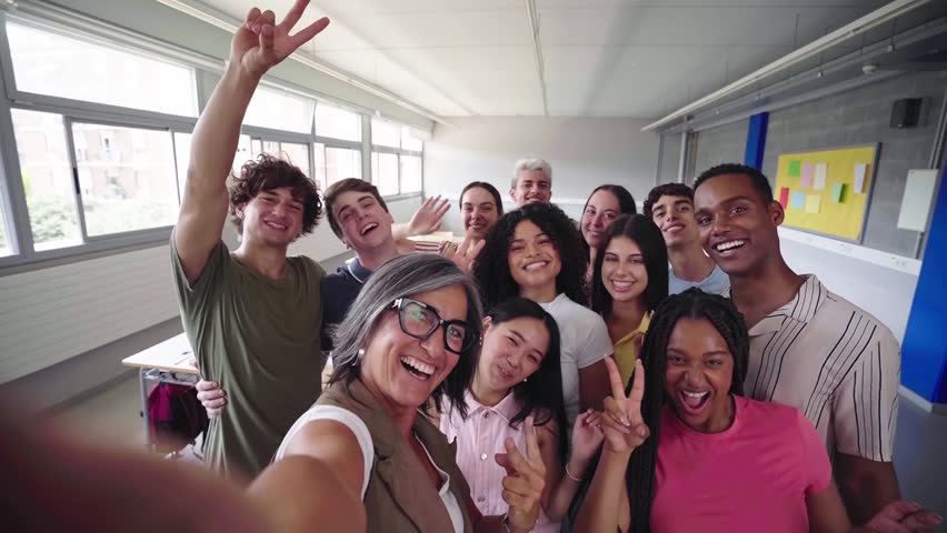 Cheerful selfie of young group of students waving and looking at camera with big smiles with their female teacher in the classroom, celebrating the back to school.