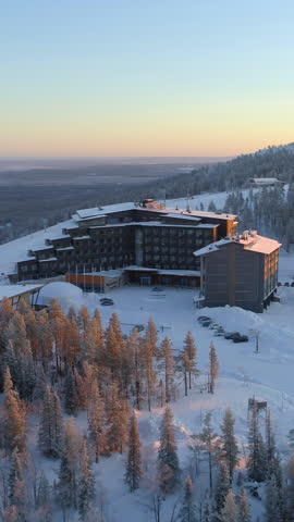 AERIAL: Flying around hilltop skiing resort surrounded by ski slopes in winter. Snowy hotel on top of ski tracks at golden winter sunset. Winter sun rising above quiet ski resort Levi in Finland