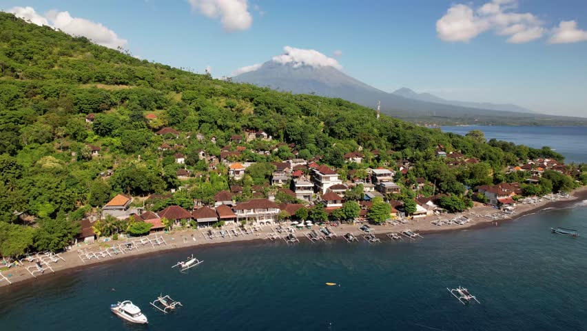 4k drone video of Jemeluk Beach with fishing boats docked on the dark sand, over the green hills covered by hotels and houses, you can see Mount Agung, an active volcano in Amed, Bali Indonesia