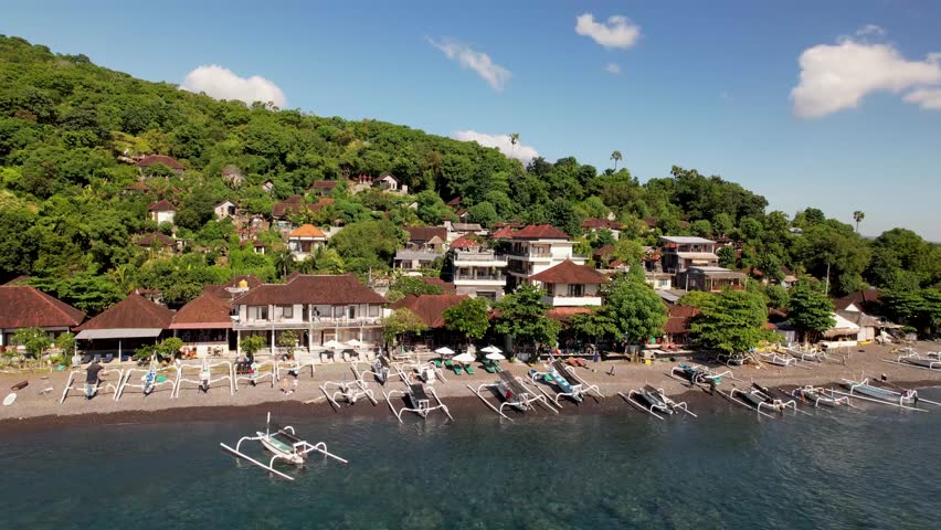4k drone video of Jemeluk Beach with fishing boats docked on the dark sand with a view over the green hills in the distance of Mount Agung, an active volcano in Amed, Bali Indonesia