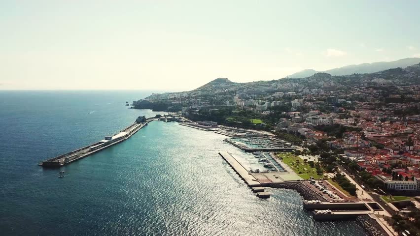 Funchal harbor in Madeira, Portugal, showcasing the marina, cityscape, and mountainous backdrop on a clear summer day, bathed in bright sunlight, drone establishing shot