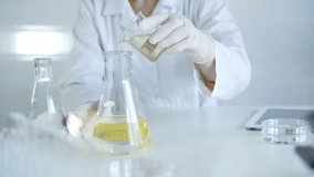 A scientist researcher, wearing white protective gloves and lab coat, is holding a beaker with yellow oil liquid while sitting at the table in laboratory, close up. Medicine and microbiology science - Powered by Shutterstock - Get 15% off with code: PIKWIZARD15