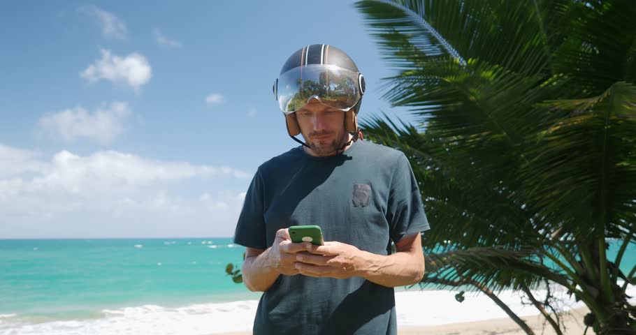 Man with moto helmet checks phone while standing near ocean and palm trees on a sunny day. 