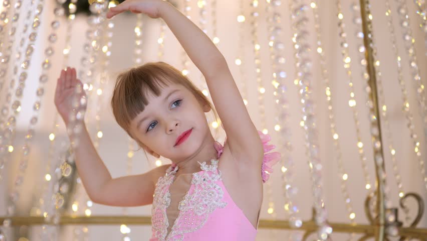 Young girl in a pink dress is dancing in front of a crystal chandelier. The scene is elegant and sophisticated, with the girl