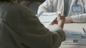 Doctor in white coat and glasses sitting at desk, taking medical records from female patient, reading them and asking questions during health consultation in clinic. Tilt-up, over the shoulder view - Powered by Shutterstock - Get 15% off with code: PIKWIZARD15