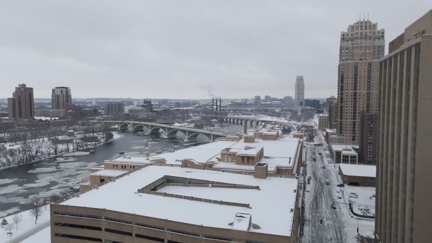 Aerial view of Minneapolis, Minnesota, showcasing snow-covered buildings, a frozen river, and the city’s iconic bridge during winter.