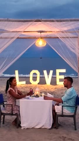A couple enjoys an intimate dinner on the beach as the sun sets, with soft lights illuminating the word love nearby. The atmosphere is filled with romance and tranquility in Thailand
