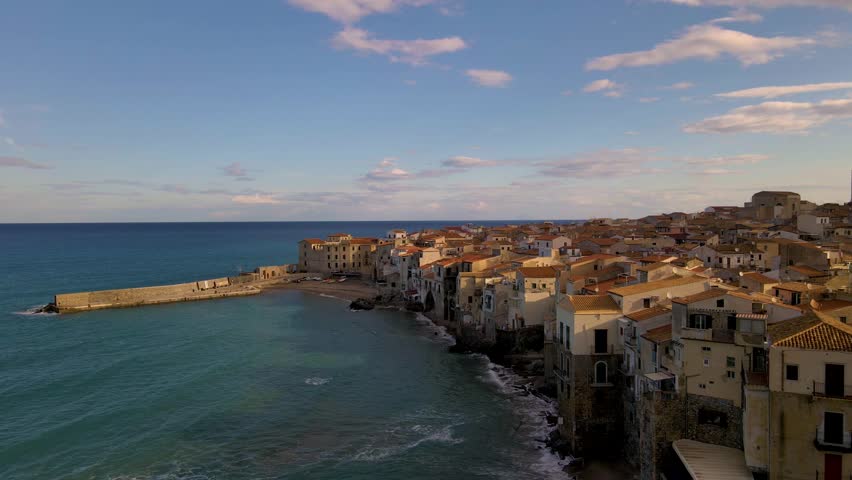 Experience the serene coastal landscape of Cefalu Sicily Italy, where picturesque houses line the shoreline against a vibrant sunset. Waves gently lap the shore, creating a tranquil atmosphere.