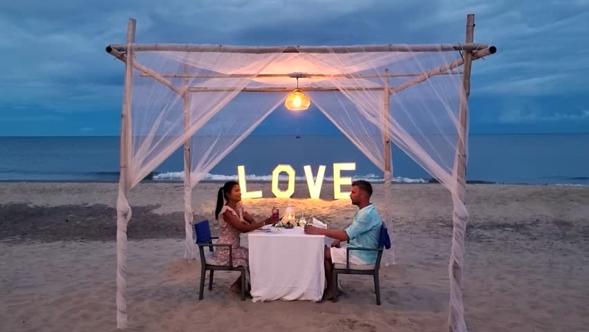Enjoy an enchanting evening on the beach in Hua Hin, Thailand, where a couple shares a candlelit dinner beneath soft lights, surrounded by serene ocean waves and a beautiful sunset.