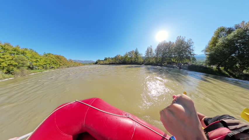 A side-angle shot of a group of rafters paddling down a wide river, wearing yellow helmets and life vests, with trees along the riverbank and a bright sun shining overhead.