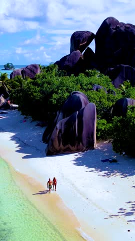 Discover the breathtaking beauty of Seychelles as two travelers stroll along the pristine beach, surrounded by lush greenery and magnificent granite boulders. Anse Source D