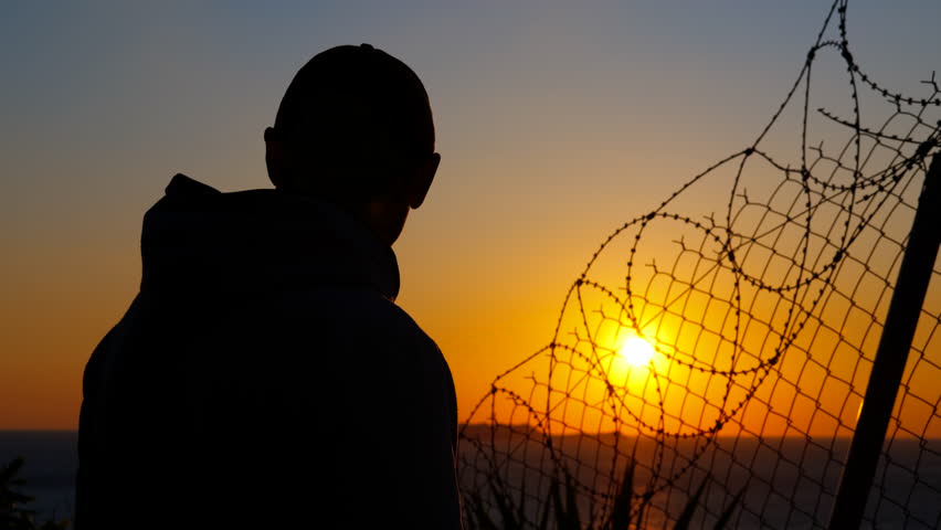 Man gazing at sunset behind barbed wire fence. Silhouette of a refugee man wearing a baseball cap looking at sunset behind a barbed wire fence, evoking themes of confinement, longing, and hope