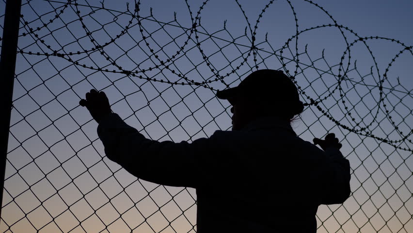 A refugee holds on to barbed wire at the border. A silhouette of a refugee woman at sunset at the border looking hopefully at the setting sun.