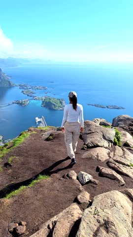 Standing on a rocky outcrop, a person enjoys panoramic views of Lofoten Norways majestic coastline, with blue waters and lush islands creating a captivating backdrop on a sunny day.