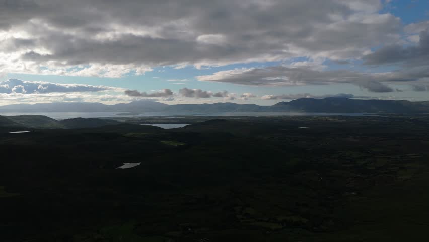 Vast green landscapes with shimmering lakes stretch out towards distant mountains, as clouds create a captivating interplay of light and shadow during late afternoon.