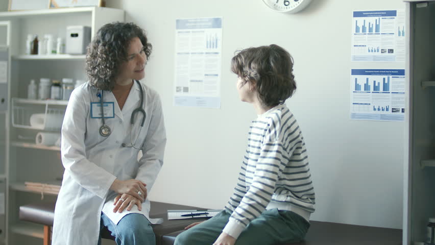 Female pediatrician wearing medical coat and stethoscope, sitting on examination table, giving handshake and attentively talking to young patient in clinic