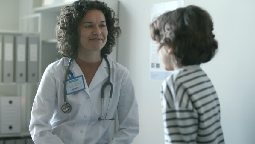 Positive female doctor in white coat with stethoscope around her neck, talking to little boy and comforting him during medical check-up at pediatric clinic