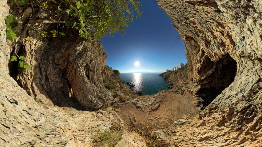 Wide Angle. A panoramic view from inside a natural rock formation overlooking the sunlit sea. The rugged cliffs and greenery frame the calm ocean, creating a beautiful and peaceful coastal scene.