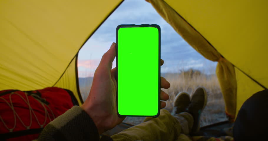 Swipe Up. Person wearing a jacket is inside a cozy tent holding mobile phone and not touching touch screen. Use green screen for copy space closeup. Chroma key mock-up on smartphone in hand.