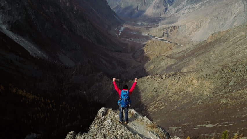 Hiker with backpack standing on mountain peak with arms raised celebrating reaching the summit enjoying scenic landscape view of autumn forest and mountains