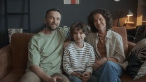 Cheerful parents and their little son sitting together on sofa in cozy living room, watching hilarious TV show, looking into camera and happily laughing in the evening at home - Powered by Shutterstock - Get 15% off with code: PIKWIZARD15