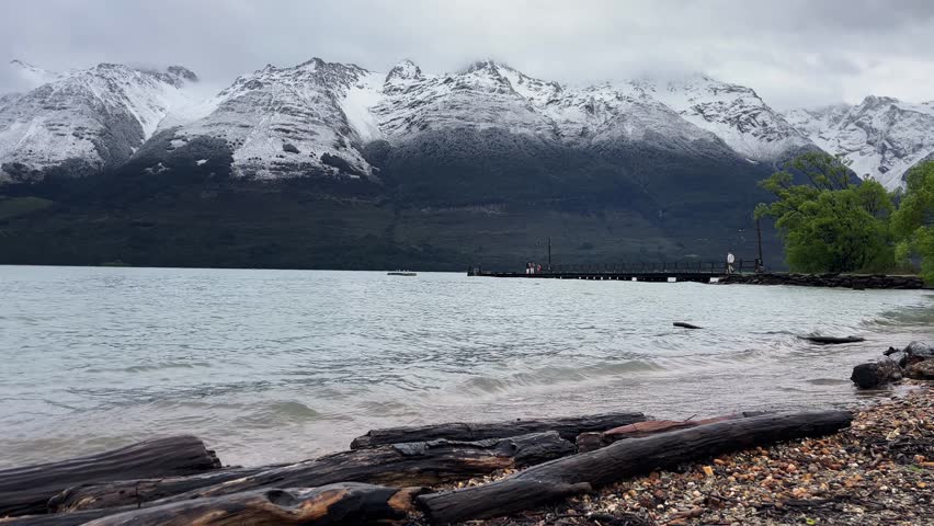 Man walks on wooden dock at Glenorchy Wharf, surrounded by lake Wakatipu and snowcapped peaks of Mount Bonpland, New Zealand.