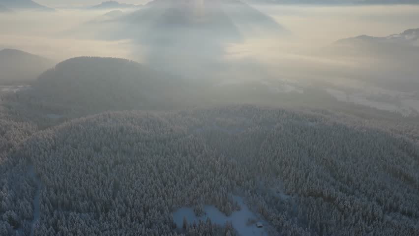 Aerial view of a breathtaking winter sunrise over snow-covered forests and misty mountains. The golden sun rises above the peaks, casting dreamy light over the foggy valley.