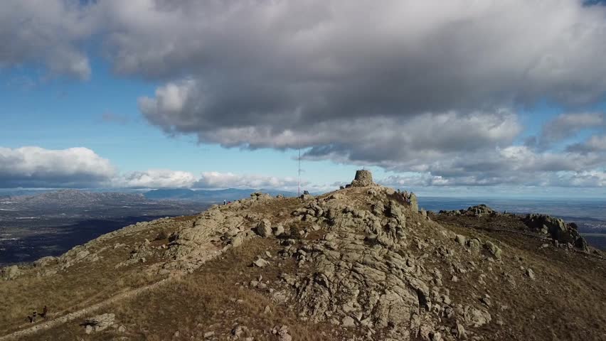 A breathtaking aerial view captures a rocky hilltop with a small structure perched on its summit. The landscape stretches out into the distance, revealing rolling hills, a distant lake.