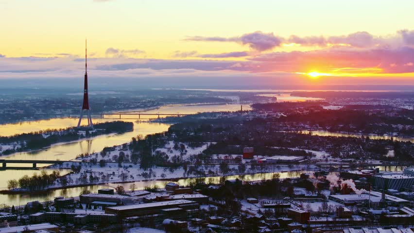 Drone glides left over Riga snowy landscape, revealing colorful winter sunset