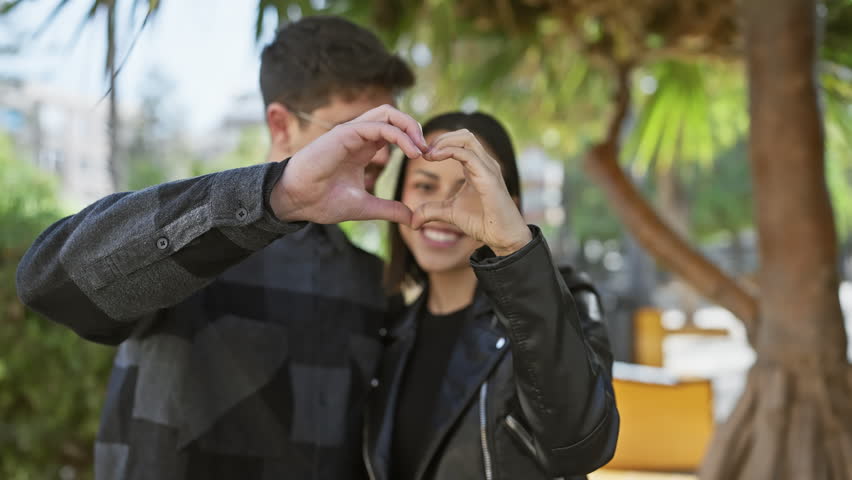 A man and woman form a heart shape with their hands on an urban street, conveying a sense of love and companionship in an outdoor setting.