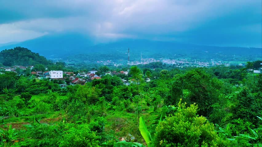 A short video of a lush green landscape with a village nestled amidst trees and rolling hills, under a cloudy sky with a mountain silhouette in the distance.
