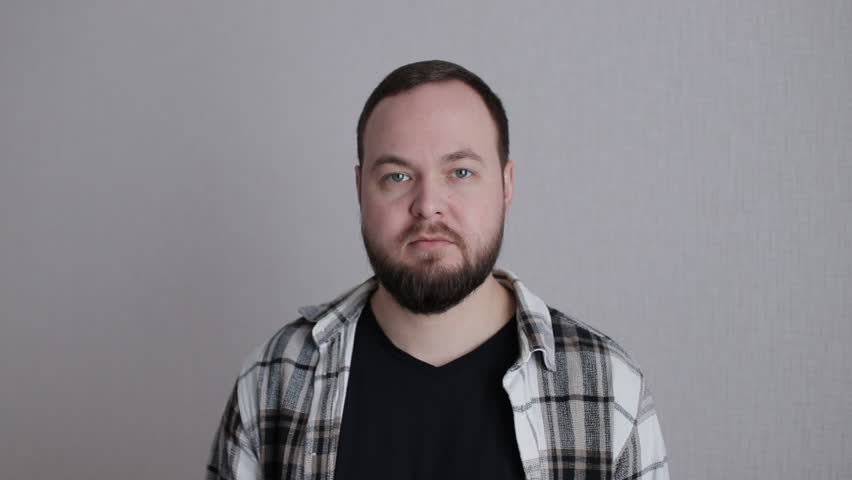 Pensive bearded male in plaid shirt contemplating, touching beard against neutral backdrop