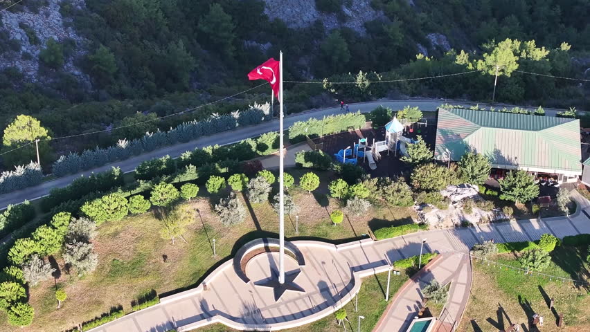 Turkish flag on a tall pole in a landscaped park with greenery and pathways near a rocky hillside, Alanya Turkey, oe01