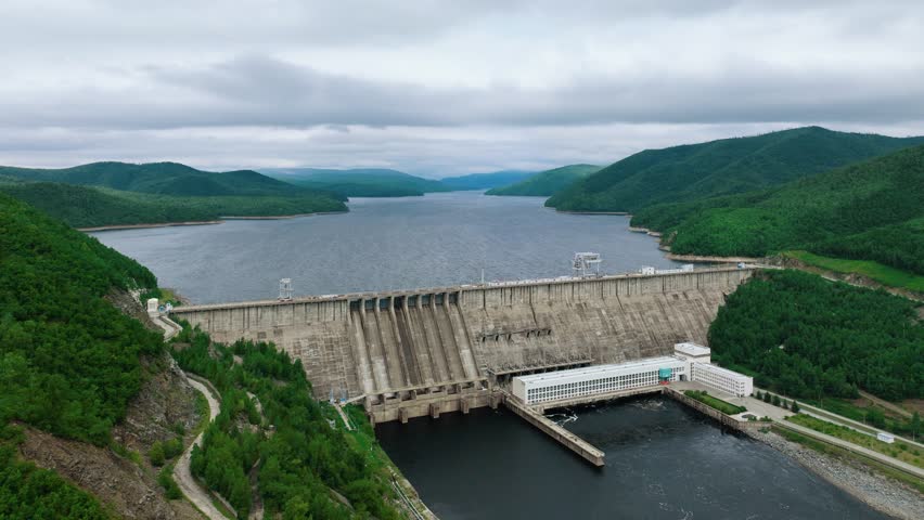 The Zeiskaya hydroelectric power station. A large dam blocks the river. Clouds are floating in the sky. The shores are overgrown with trees. Electricity is being generated.