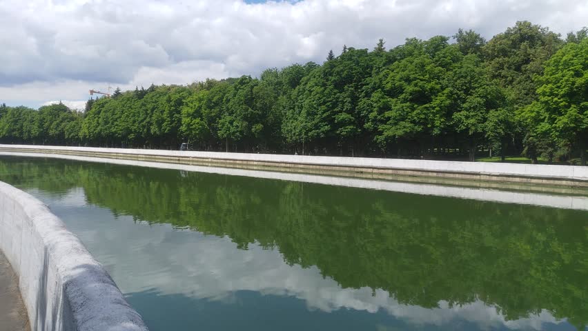 A river with concreted banks and parapets flows through the park. The water reflects trees and blue sky with clouds. Rides a tractor on the embankment. On the opposite bank are trees and a crane