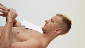 Vertical video. Hand care. Skin treatment. Cosmetic product. Profile of shirtless man with white towel on shoulder applying moisturizing lotion from tube isolated on white. - Powered by Shutterstock - Get 15% off with code: PIKWIZARD15
