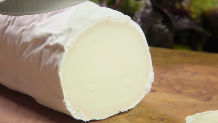 The knife cuts off a piece of Sainte maure de Touraine goat cheese on a wooden board on a background of fresh vegetables. High quality 4k footage