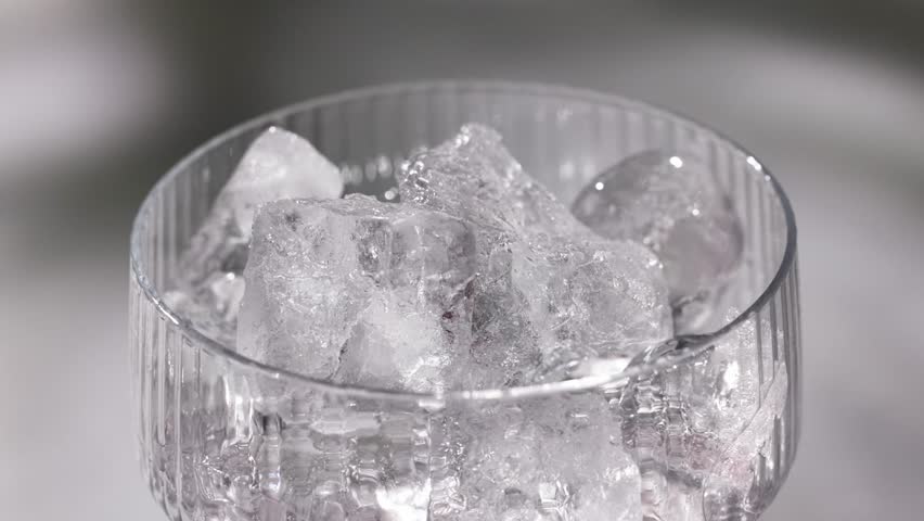 fresh red drink pouring into a cocktail glass with ice cubes on a gray sunny restaurant tabletop, slow motion, zoom in