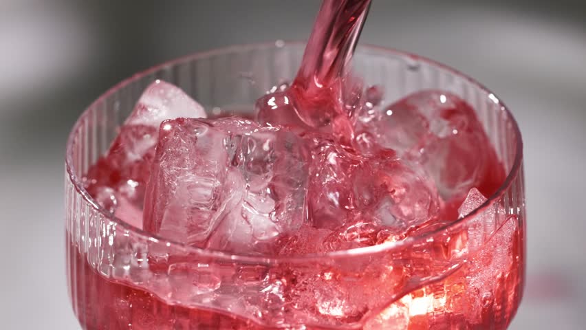 fresh red drink pouring into a cocktail glass with ice cubes on a gray sunny restaurant tabletop, slow motion, zoom in