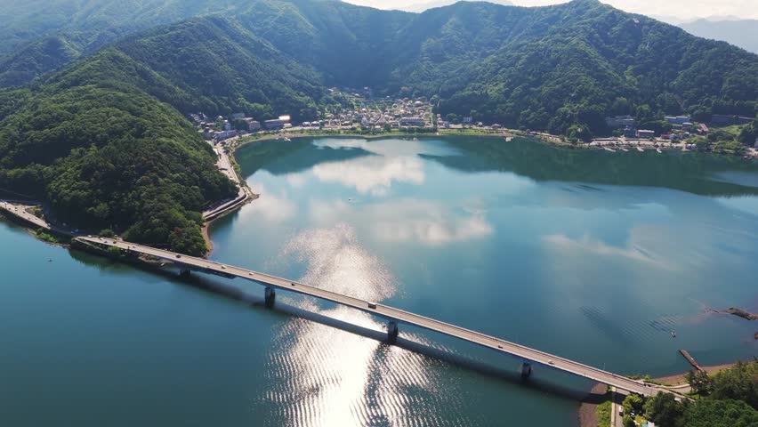 Aerial view of a long bridge crossing lake Kawaguchiko, surrounded by green mountains. The calm blue water reflects the sky. A peaceful and scenic location in Japan.